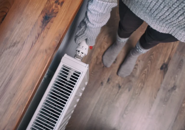 Person Wearing A Gray Sweater And Gloves Adjusting A Radiator Thermostat, Standing On A Wooden Floor.