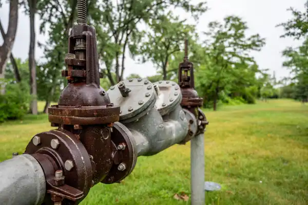 Metal water pipe equipped with valves and flanges sits in a grassy park area, surrounded by trees, ready for backflow testing.
