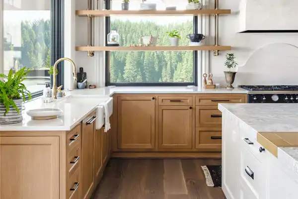 A modern kitchen with light wood cabinets, white countertops, open shelves adorned with greenery, and a large window offering a serene forest view. The gleaming stainless steel faucet adds a sleek touch, seamlessly combining style with easy access for any future plumbing repair needs.