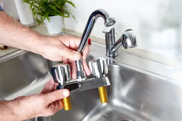 A person expertly handling kitchen plumbing, either installing or removing a faucet over the sink. Visible plumbing parts align perfectly as a potted plant adds a touch of greenery in the background, creating a harmonious kitchen install scene.