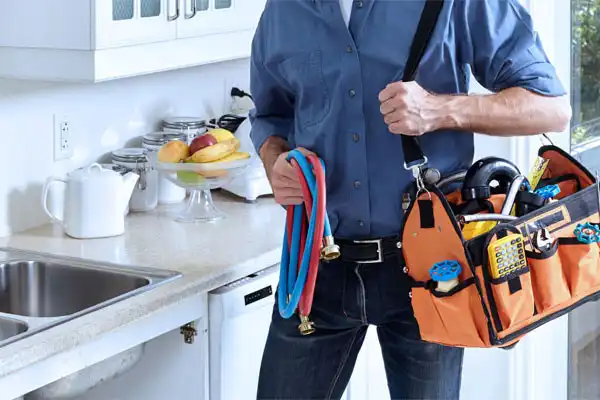 A person in a blue shirt, ready for kitchen plumbing repair, stands by the sink with a tool bag and hoses, near a fruit bowl.