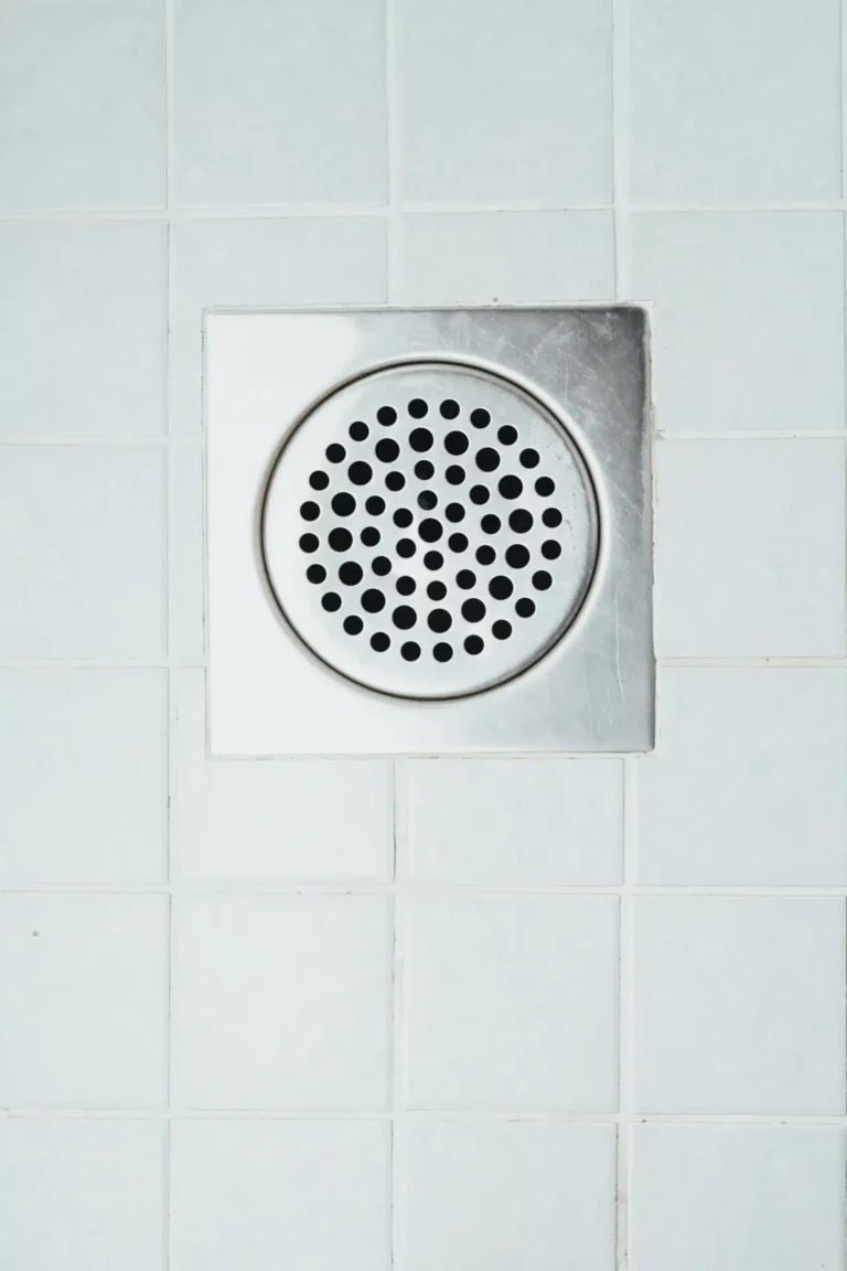 A square metal shower drain cover with circular perforations is set in a white tiled floor, ensuring efficient water flow that's perfect even when rooter service might be needed.