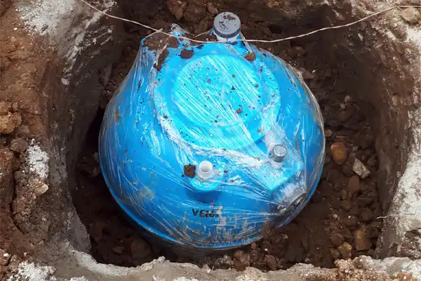 A blue plastic container, resembling a mini septic tank install, is partially buried in a dirt hole. It's covered in clear plastic wrap and surrounded by soil and stones.