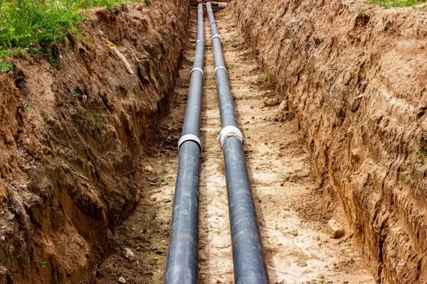 Two large black pipes, part of a sewer line install, lie in a narrow trench dug in the ground, surrounded by dirt.