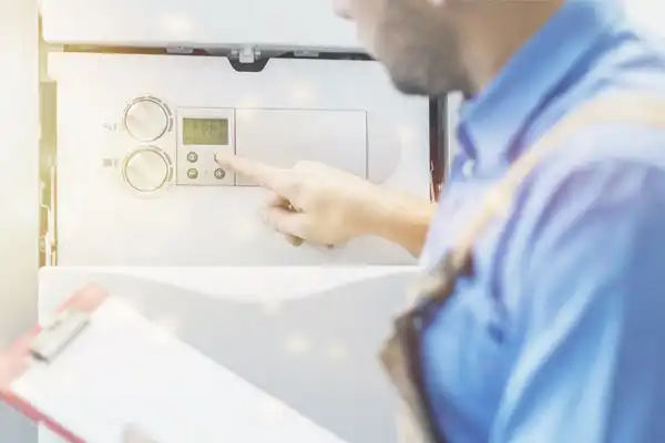 A person in blue overalls adjusts the tankless water heater settings on a control panel, holding a clipboard.