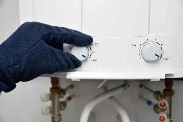 A gloved hand precisely adjusts a dial on a sleek white tankless water heater, with visible pipes and control knobs ready for its tune-up.