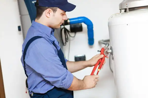 A person in a cap and overalls uses a wrench to adjust a pipe on a large white appliance during a water heater tune-up.