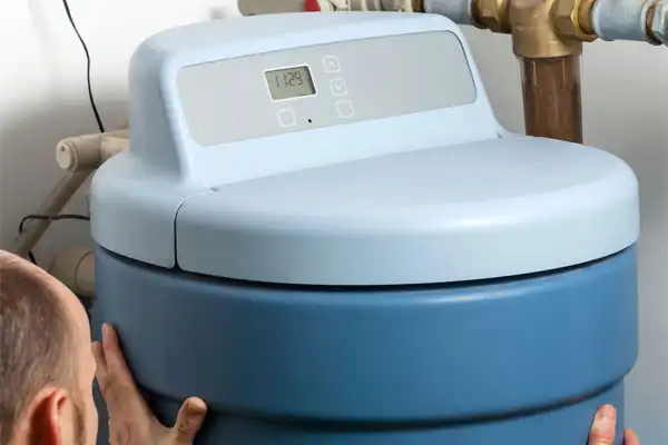 A person carefully adjusts a large blue water softener with a digital control panel on top, its installation complete, surrounded by an intricate network of pipes.