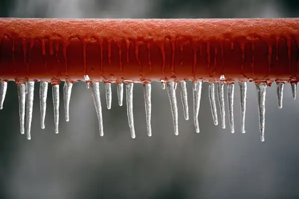 Icicles hanging from a red pipe on a cold day reveal the aftermath of a frozen burst pipe.