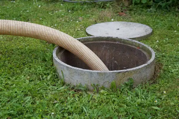 A large, corrugated hose is inserted into an open septic tank surrounded by grass for septic tank pumping. The concrete lid is partially removed and placed nearby.