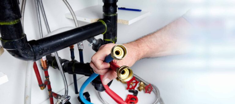 A person is holding blue and red hoses near black pipes and valves, possibly working on a plumbing install or maintenance in a utility area.