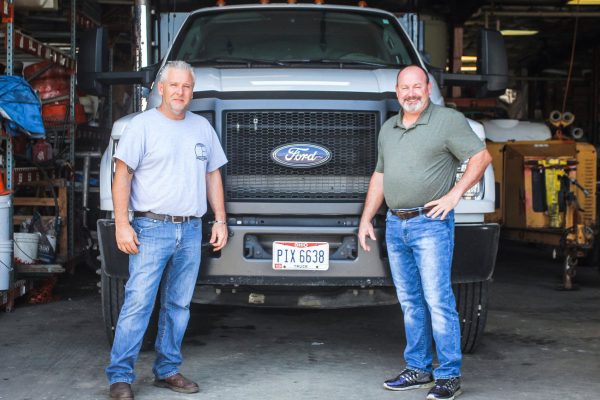 Two Men Stand In Front Of A Large Ford Truck Inside A Garage. Both Are Casually Dressed In Jeans And T-Shirts, Showcasing The Laid-Back Spirit Our &Quot;About Us&Quot; Team Embodies.