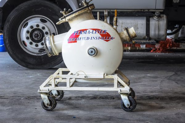 In A Garage Setting, A White Perma-Liner Certified Installer Machine Sits On Wheels, With A Vehicle In The Background. Discover More About Us And Our Commitment To Quality Installations That Set Industry Standards.